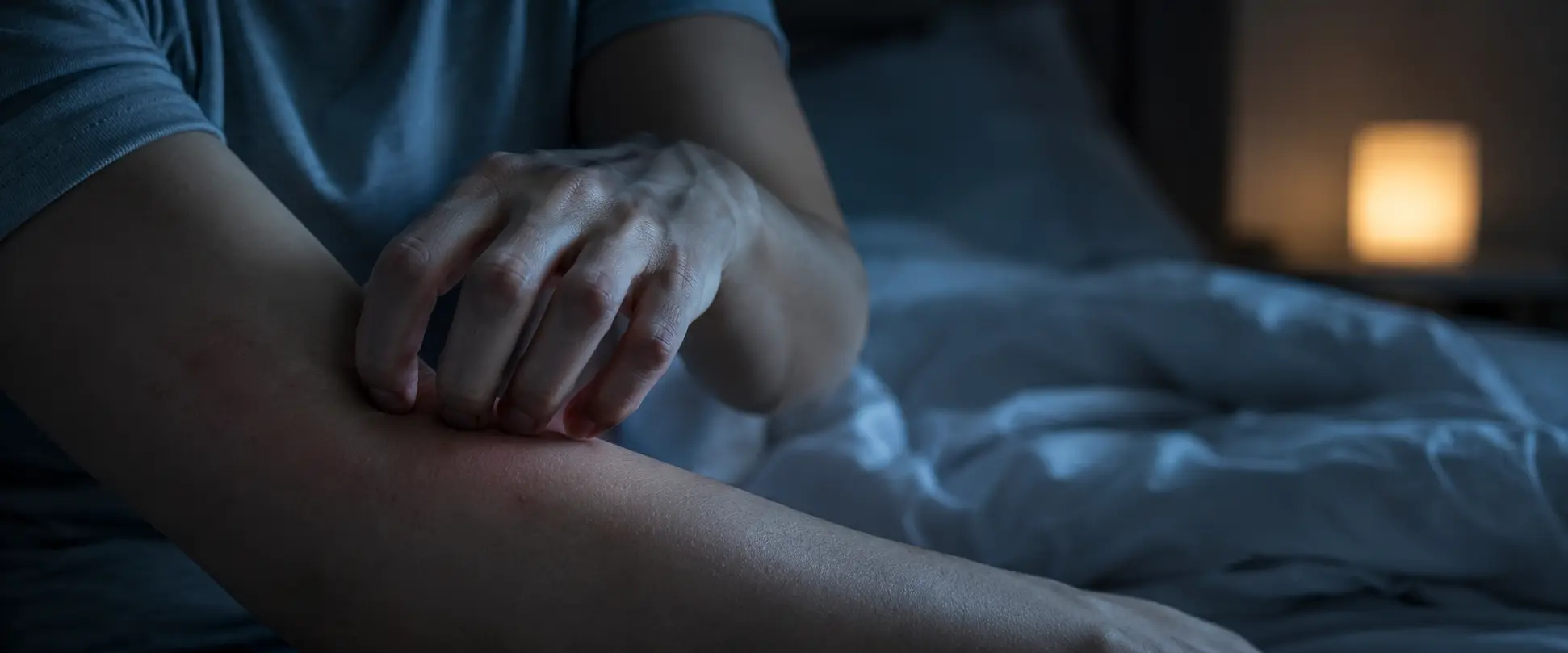 Person scratching their forearm due to nighttime itching in bed, with out-of-focus sheets and a night light.